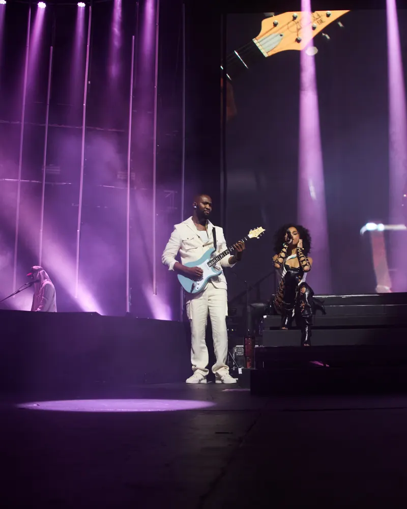 Iyke M Abu in a white suit playing a Stratocaster on an arena stage under purple lights