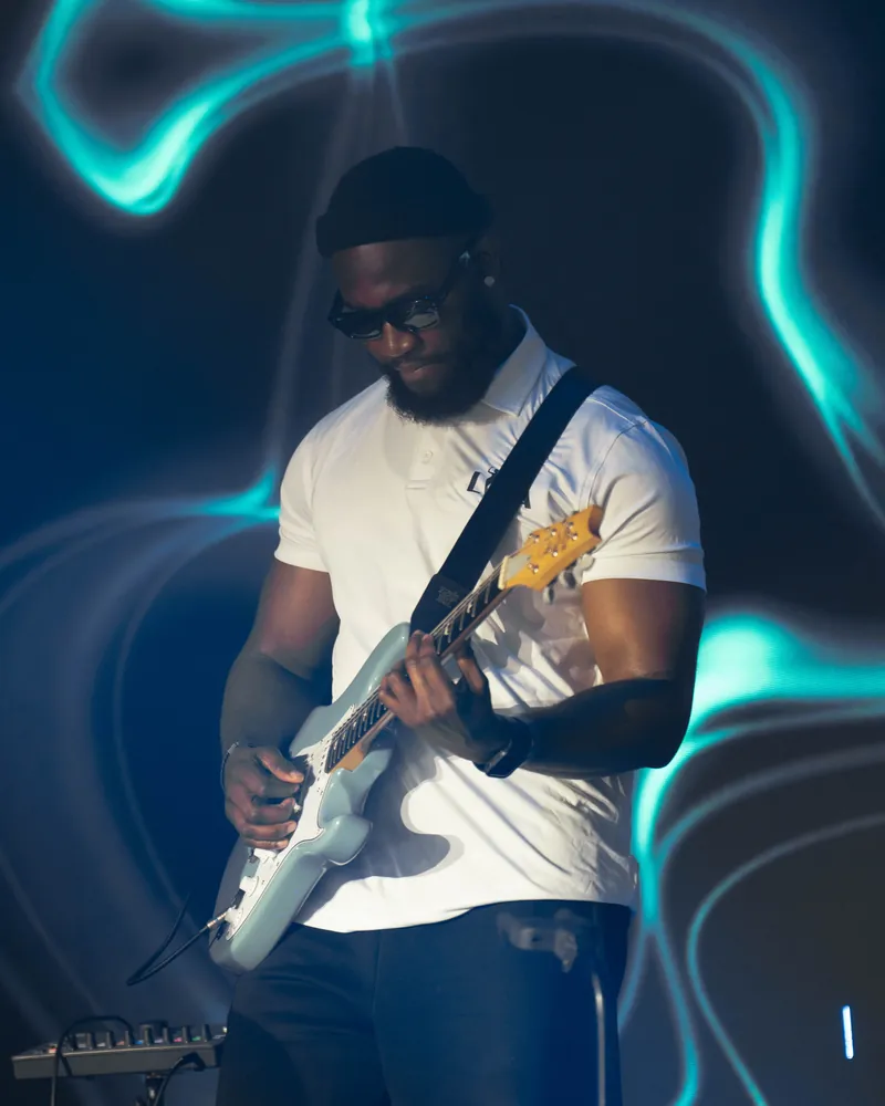 Iyke M Abu in a white T-shirt and sunglasses playing a white Stratocaster against a neon stage backdrop