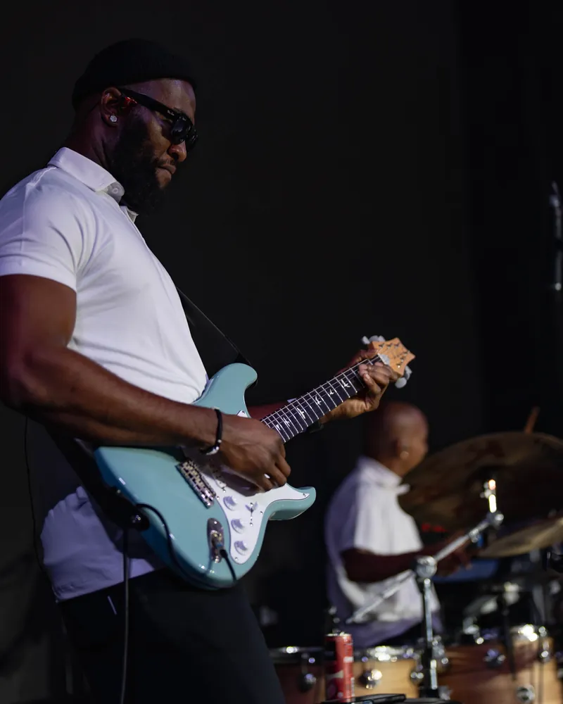 Iyke M Abu playing a light-blue Stratocaster on stage, with the drummer behind him
